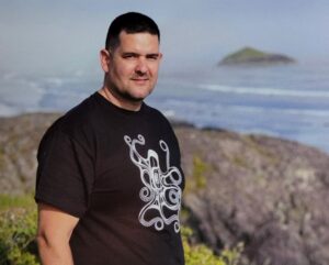 Photo of Levi Purjue, wearing a black t-shirt, with a rocky shore and ocean behind him.